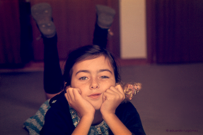 Niña con la Barbie tumbada en el suelo y los pies al aire, mirando a cámara.