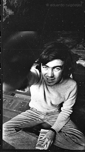 Chico sentado mirando hacia arriba a la cámara. En una mano lleva una caja de pinturas de cera.