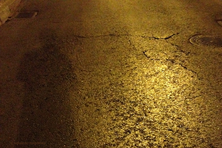 Sombra de un paseante por la noche en una calle. A la derecha se ve una alcantarilla.