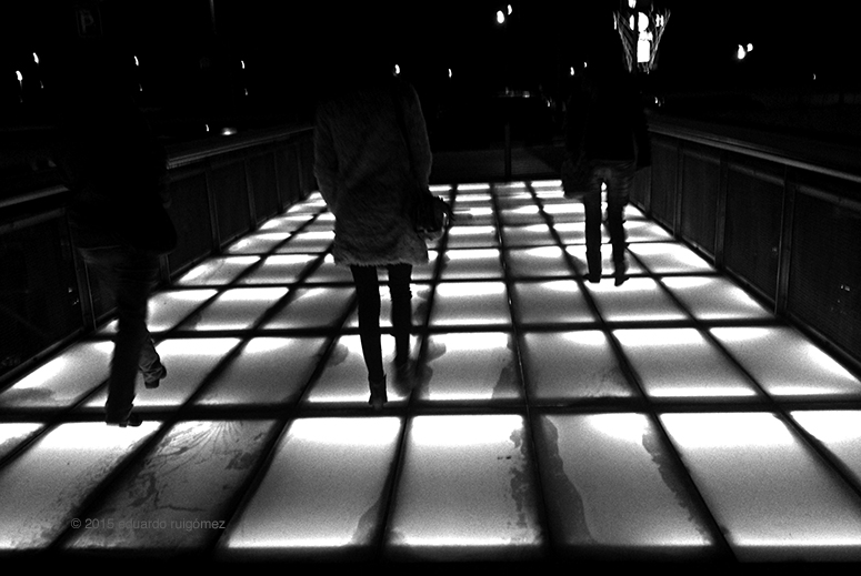 Tres personas caminan en la noche sobre un puente de cristal iluminado.